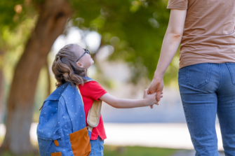 An early years child wearing a back pack and holding hands with an adult. They are looking up at them.  