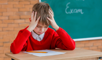Primary school pupil in white polo shirt and red jumper sits at a desk with paper and pencil in front of them. They have their head in their hands and they are looking down.