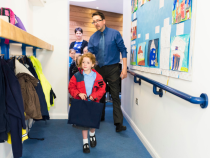 A primary aged pupil walks into the school corridor with their book bag and coat. A teacher is walking closely behind them.