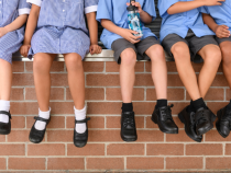 A group of primary school pupils sit on a wall in a row. Their legs dangle over the edge and you can see they shiny leather school shoes.